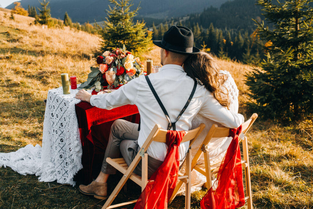 An attractive couple of newlyweds, a tender moment before the kiss. Man and woman in festive attire sitting at table near wedding arch in boho style. Wedding outdoors.